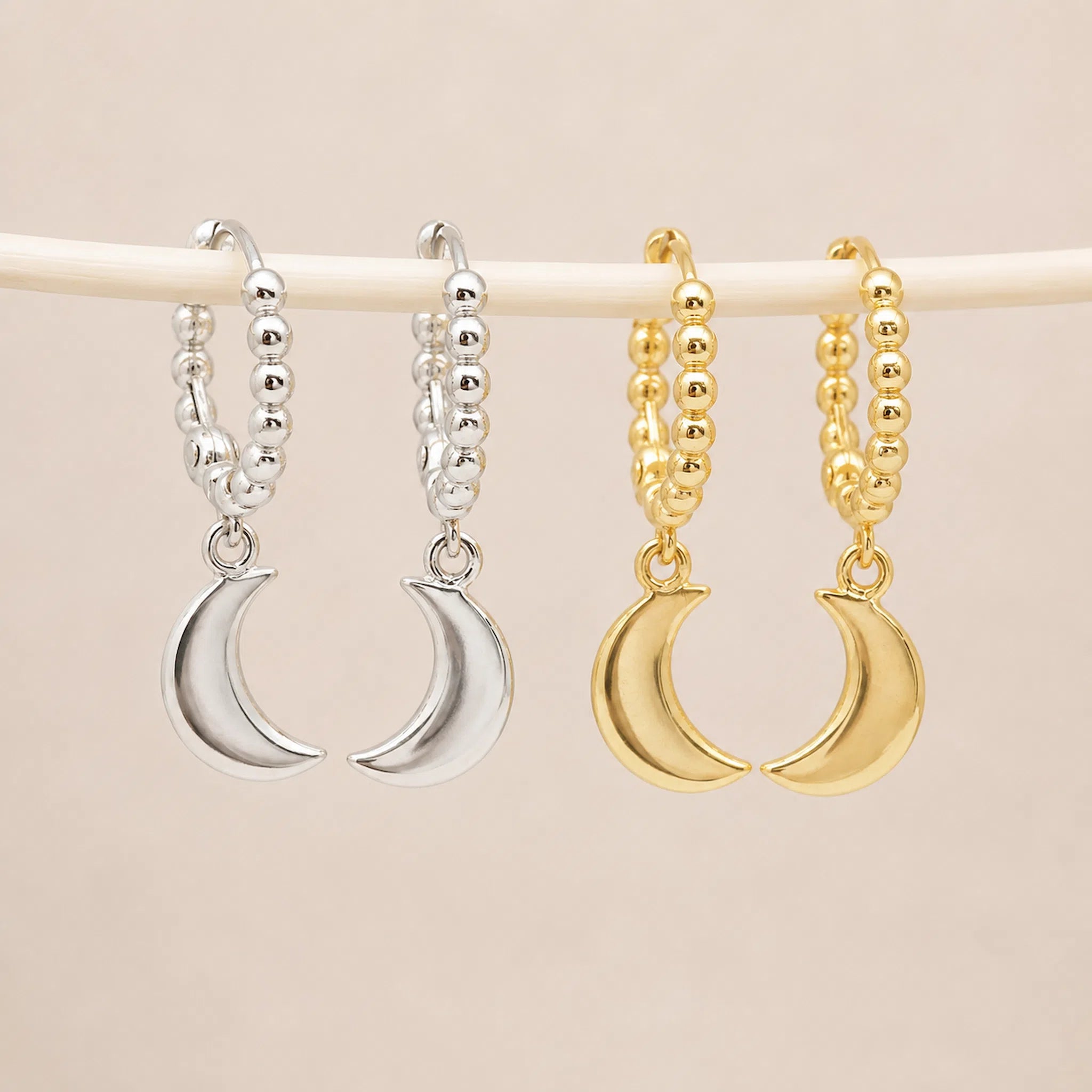 Two pairs of hoop earrings with crescent moon charms hang from a white rod against a light beige background. The left pair is silver, and the right pair is gold.