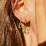 A close-up of a person's ear wearing two small gold earrings: one with a crescent moon charm and one with a star charm, against warm sunlight.