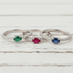 Three silver rings with colored gemstones on a wooden surface with flowers in the background