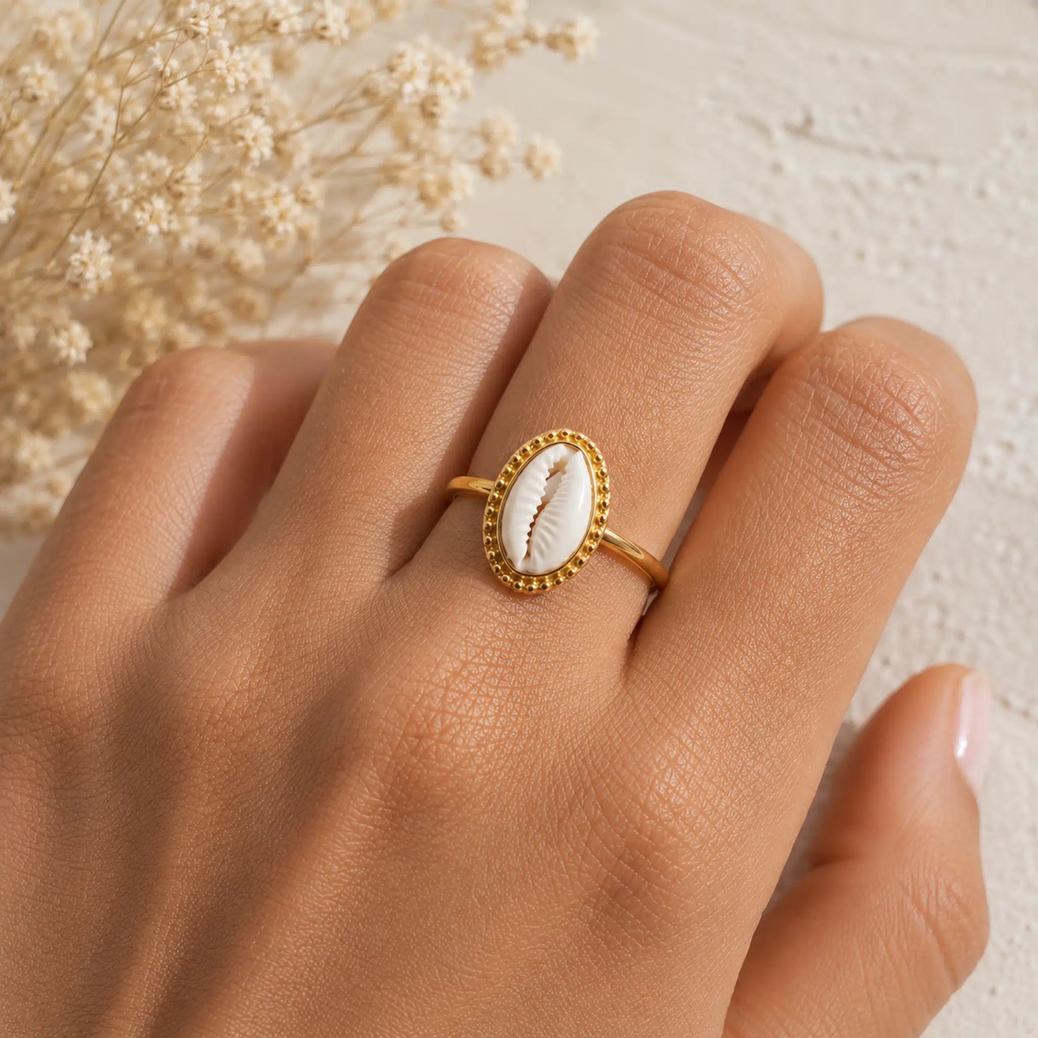 A gold ring featuring an oval white cowrie shell inlay, worn on a person's finger against a light textured background with dried flowers.