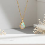 Gold necklace with a teardrop-shaped opal pendant on a white surface with dried flowers.