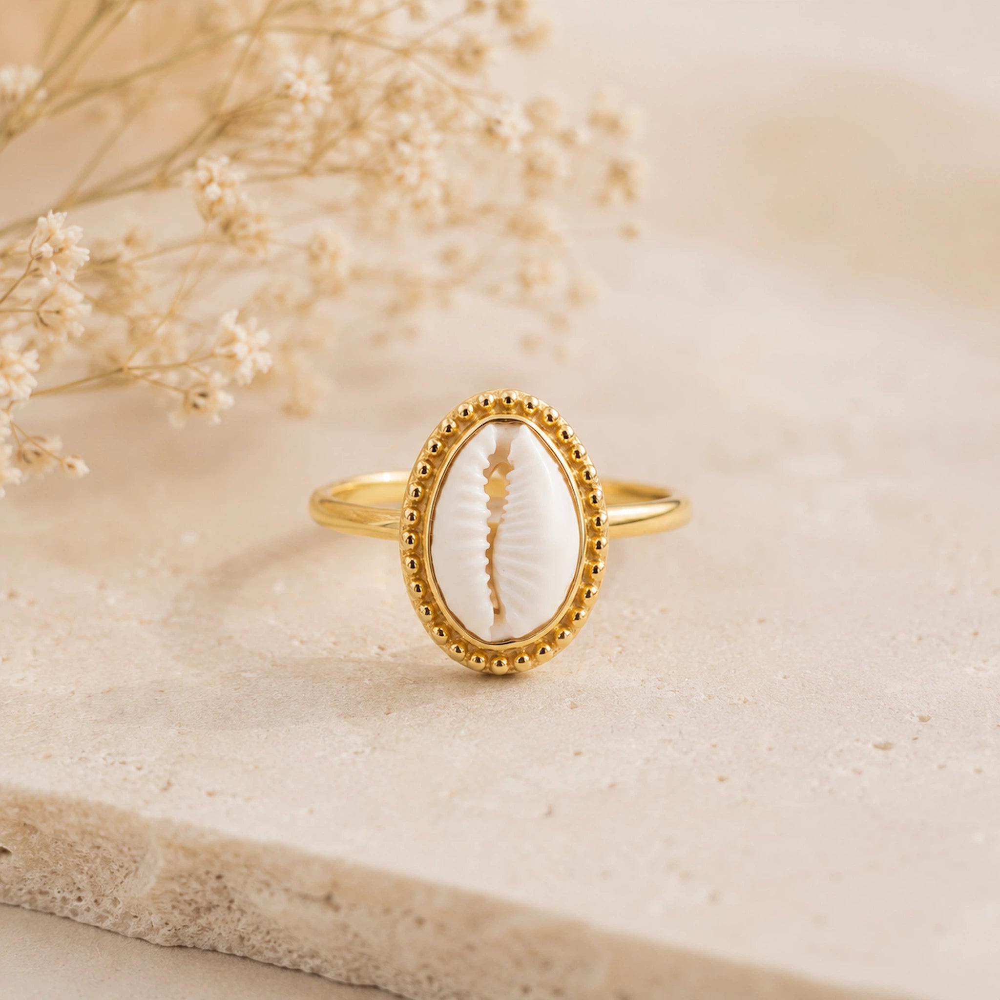A gold ring featuring an oval natural cowrie shell centerpiece, displayed on a textured beige stone slab with dried baby's breath flowers in the background.