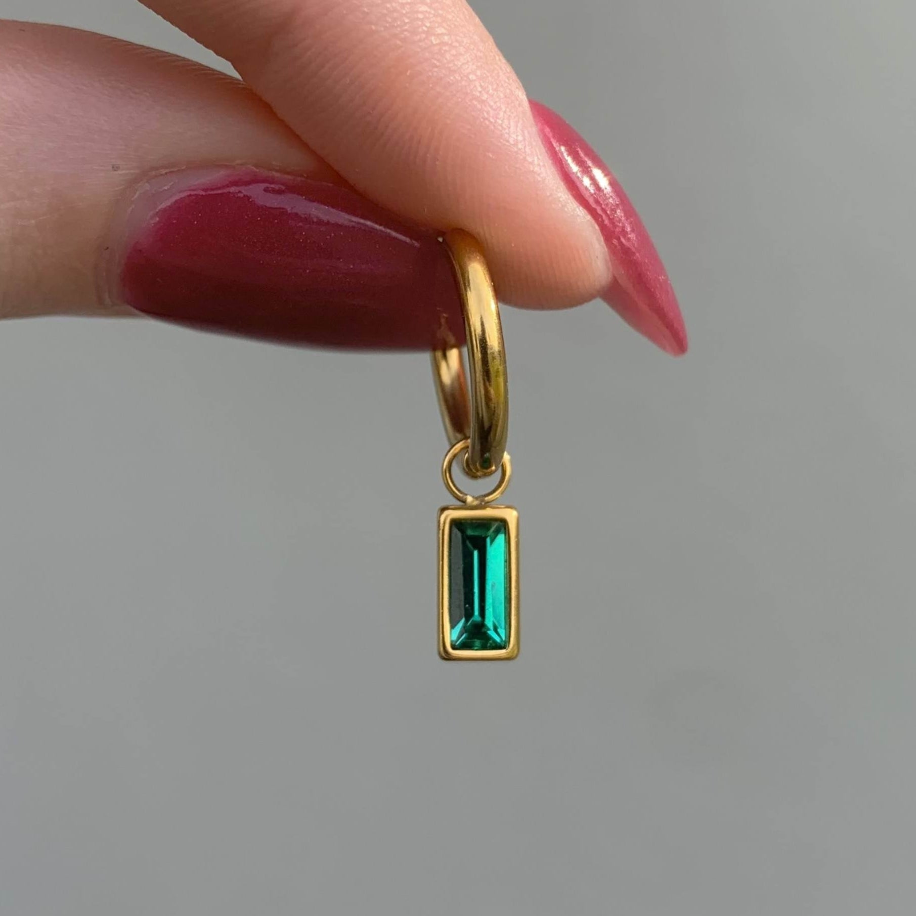 Gold hoop earring with a green gemstone held between fingers against a neutral background