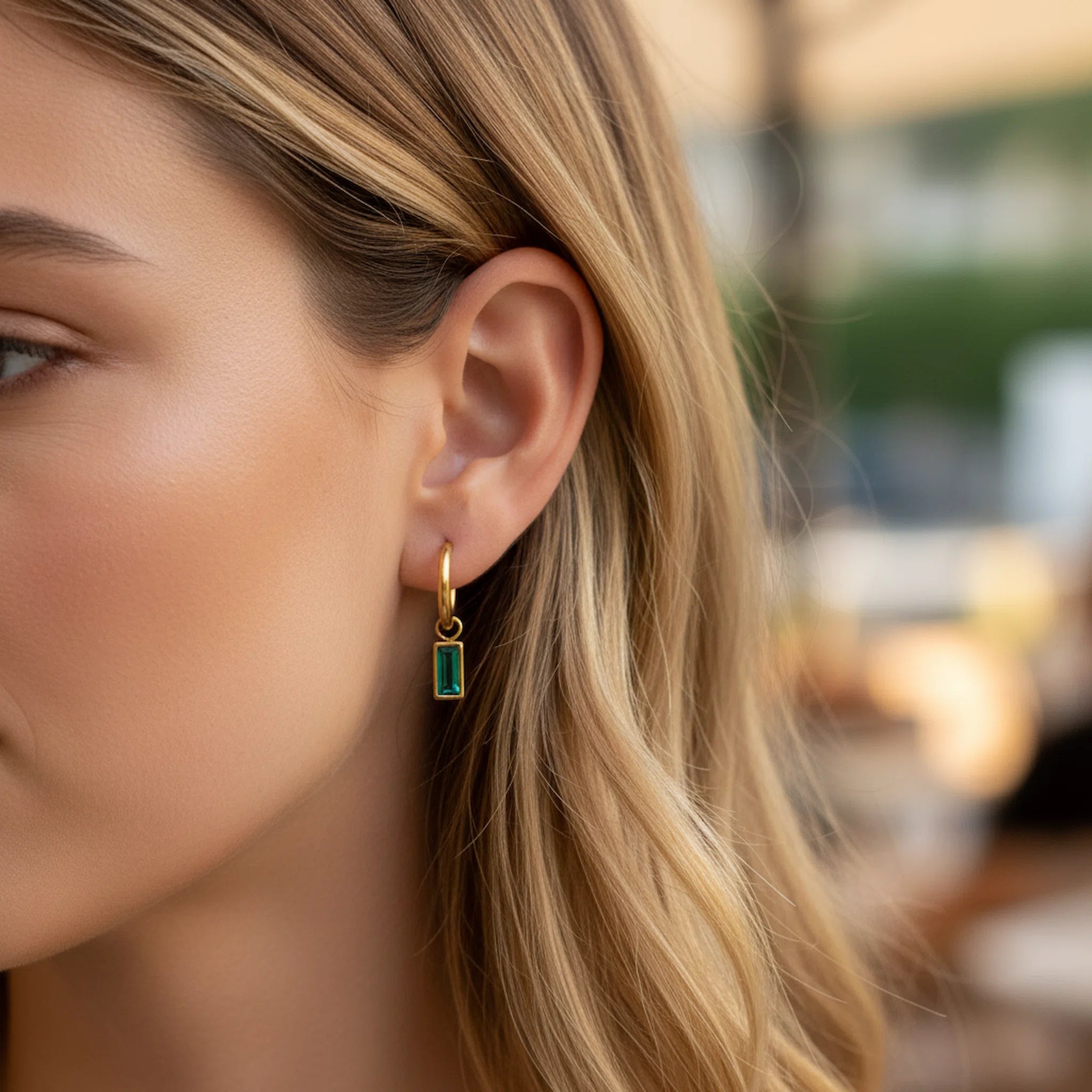 A close-up of a woman's ear wearing a delicate gold huggie hoop earring with a small rectangular green gemstone pendant, set against a softly blurred outdoor background.