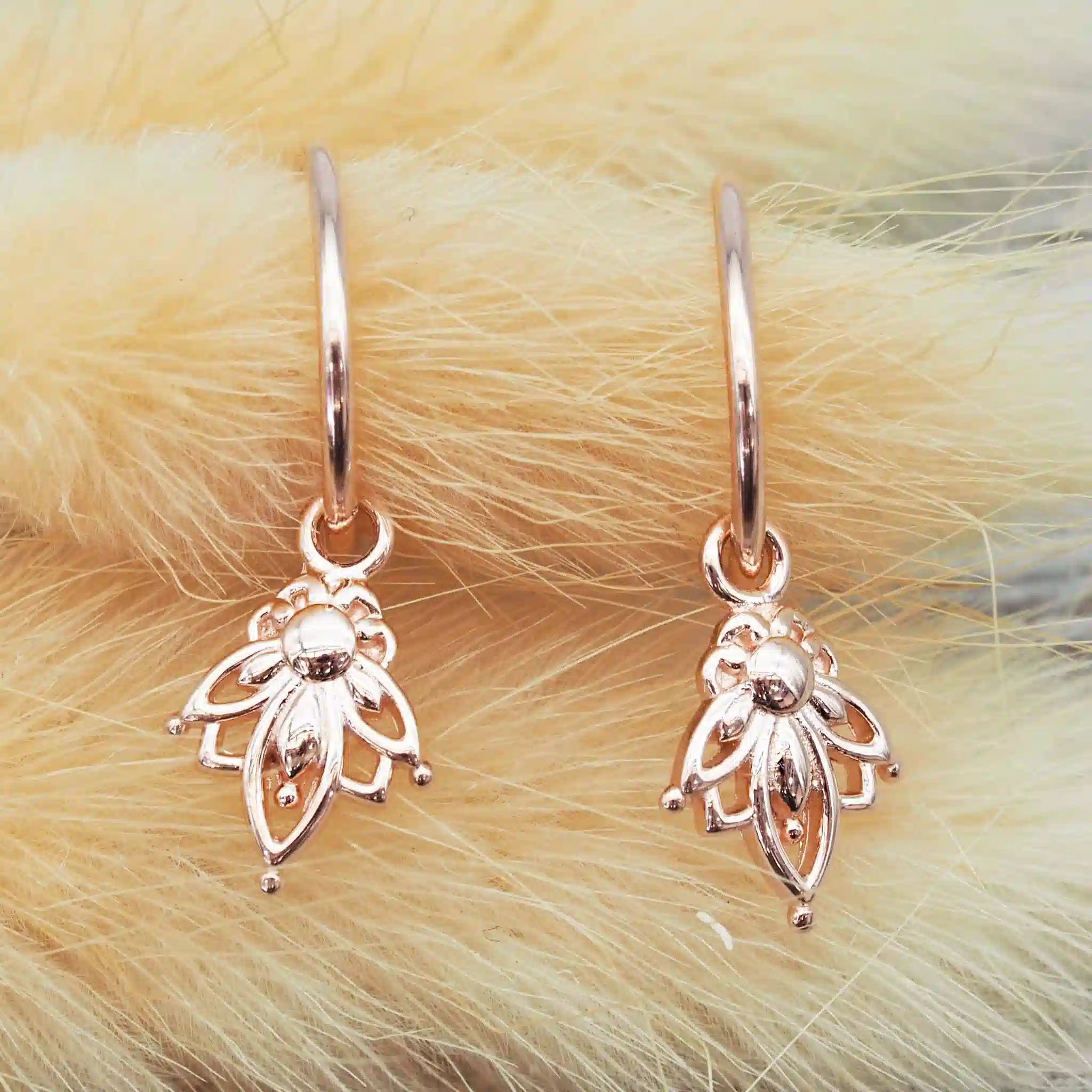 Pair of rose gold lotus hoop earrings with lotus flower charms on a fluffy beige background