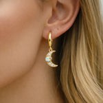 Gold hoop earring with a moon pendant with opal and cubic zirconias worn by a person with blonde hair.