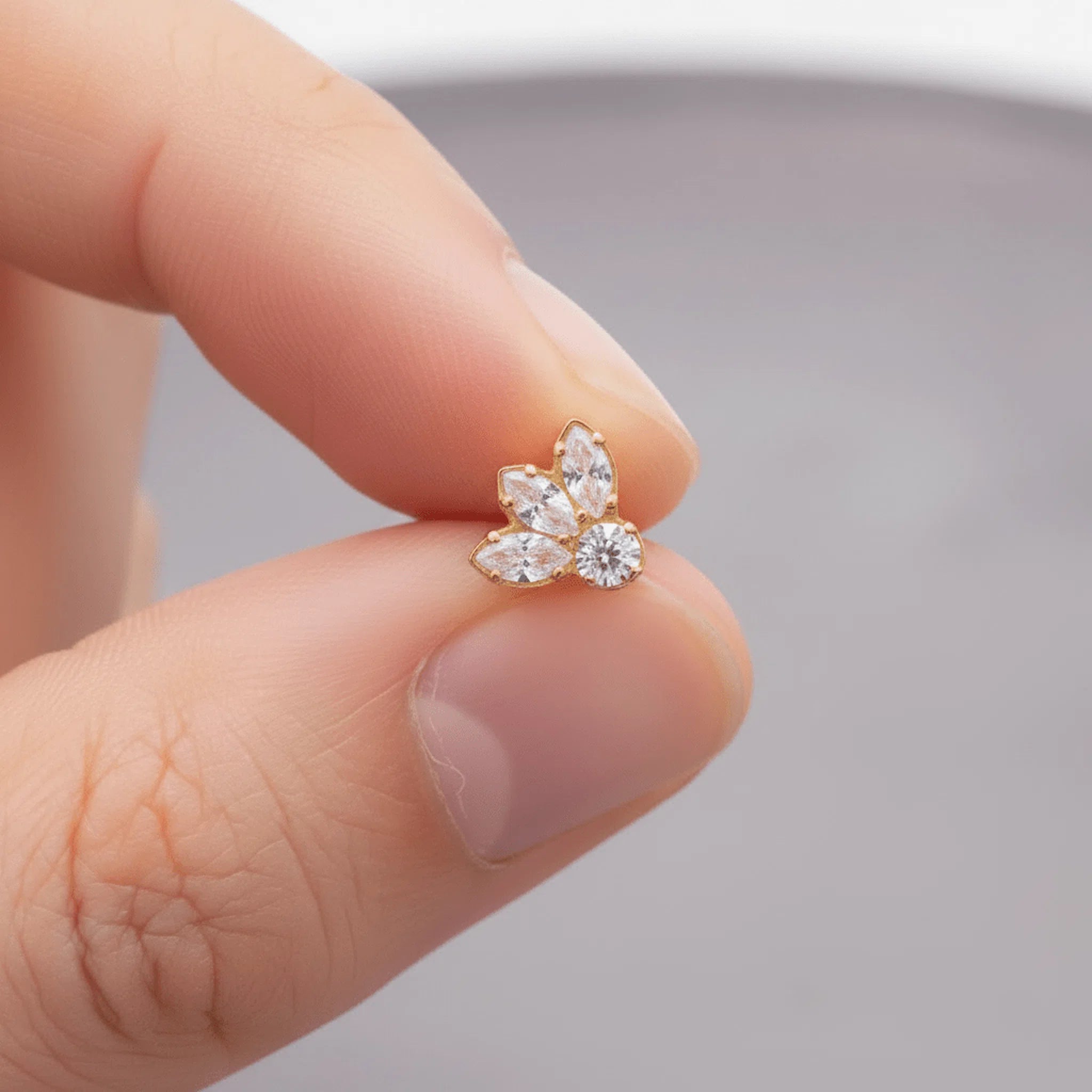 A tiny gold flower-shaped stud earring with clear gemstones, held between two fingers to show its small scale.