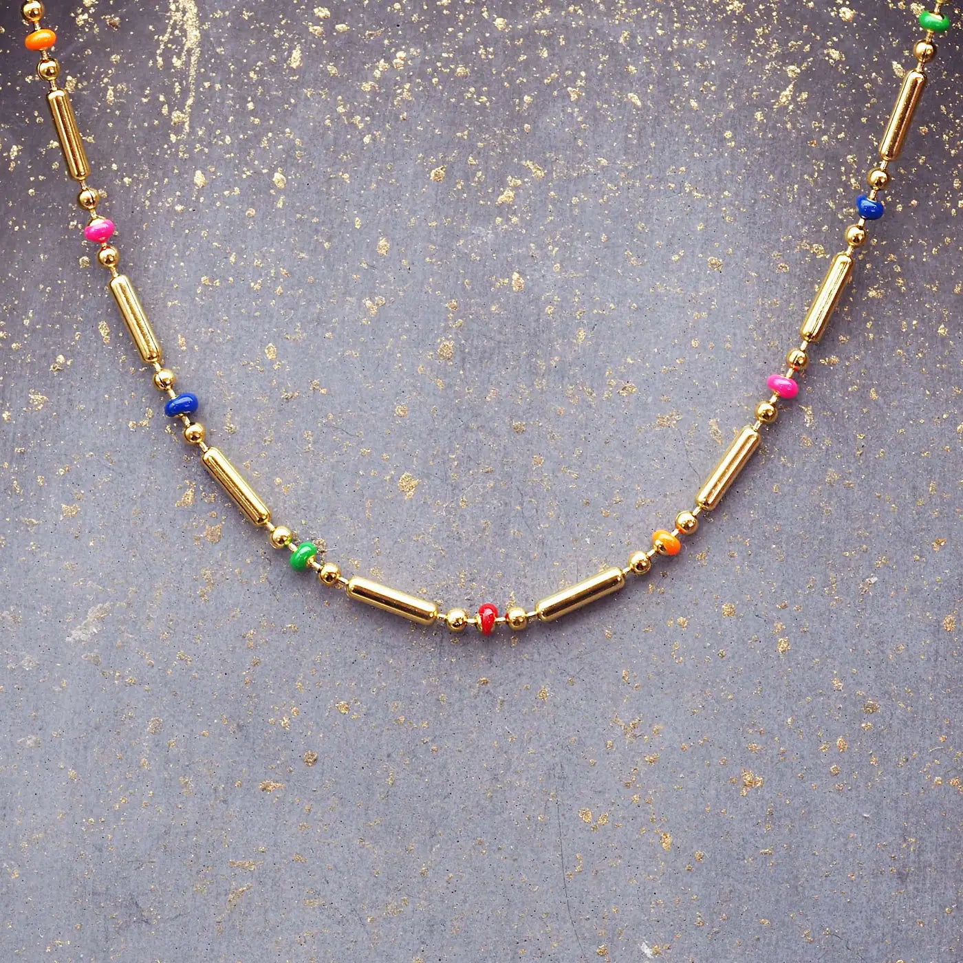 Dainty gold beaded necklace with colourful rainbow seed beads on a textured neutral background – Indie and Harper jewellery.