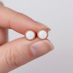 A close-up of a person's fingers holding a pair of round stud earrings with iridescent white opal-like centers in rose gold bezels against a soft light gray background.