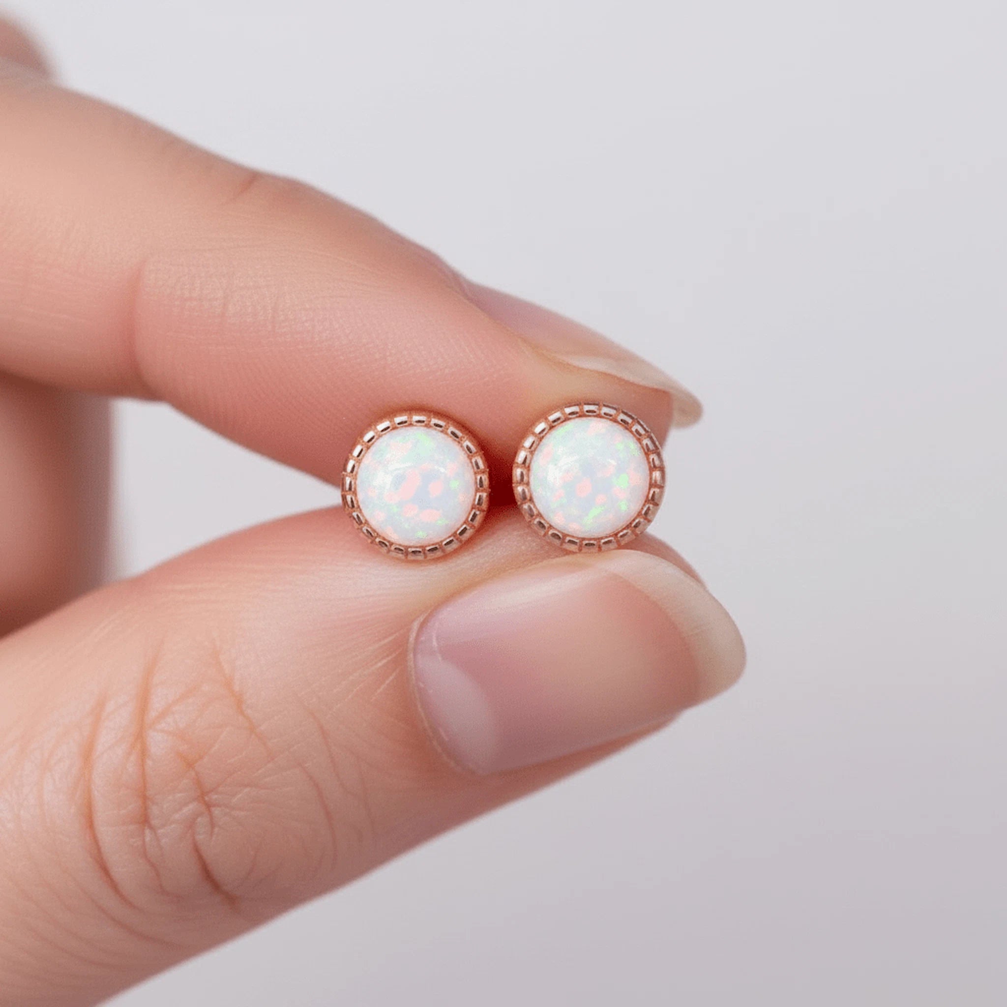 A close-up of a person's fingers holding a pair of round stud earrings with iridescent white opal-like centers in rose gold bezels against a soft light gray background.