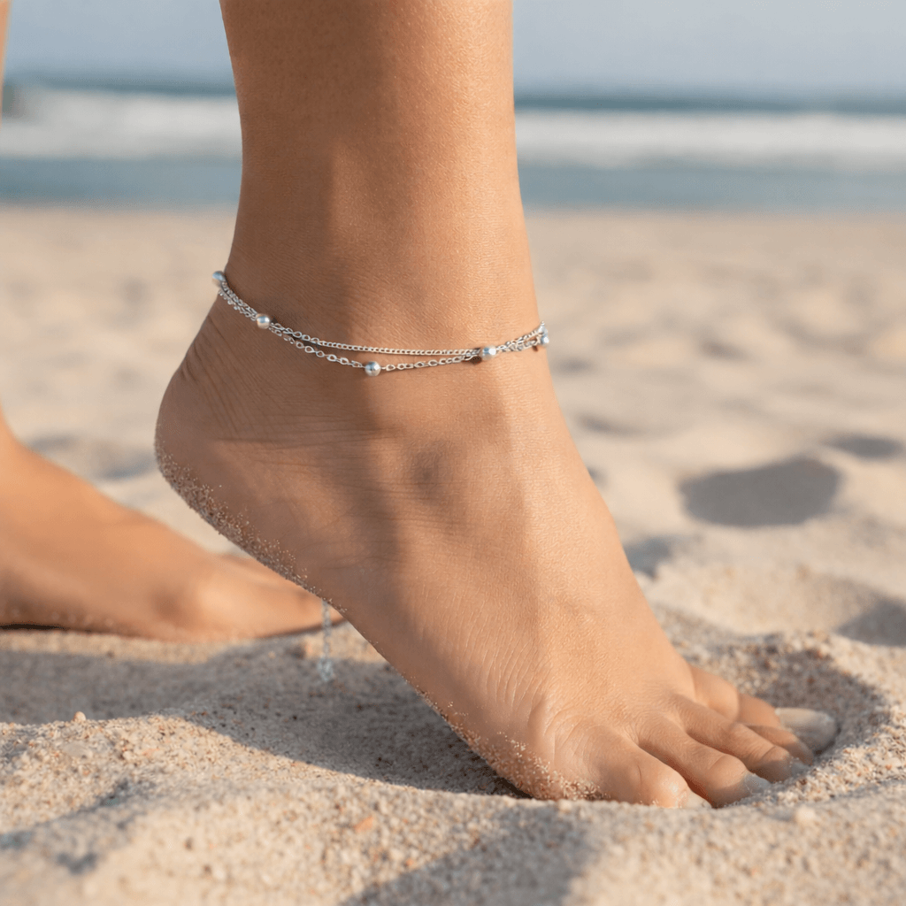 Foot wearing a silver anklet on a sandy beach with ocean in the background