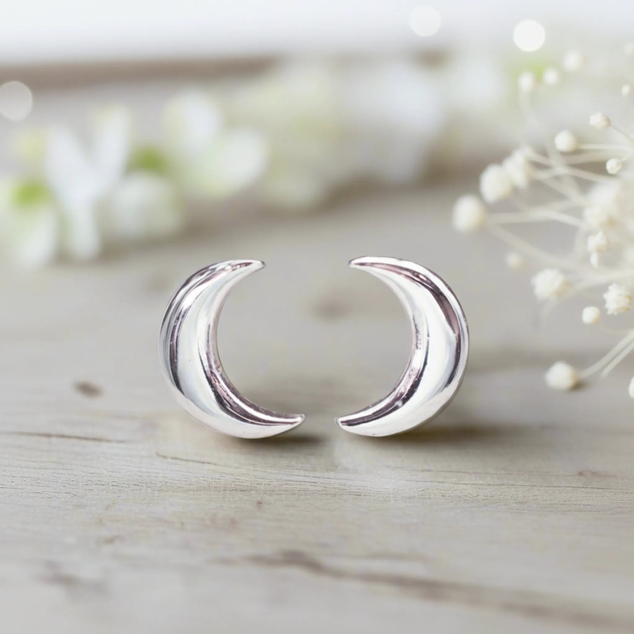 Silver crescent moon earrings on a light wooden surface with blurred flowers in the background