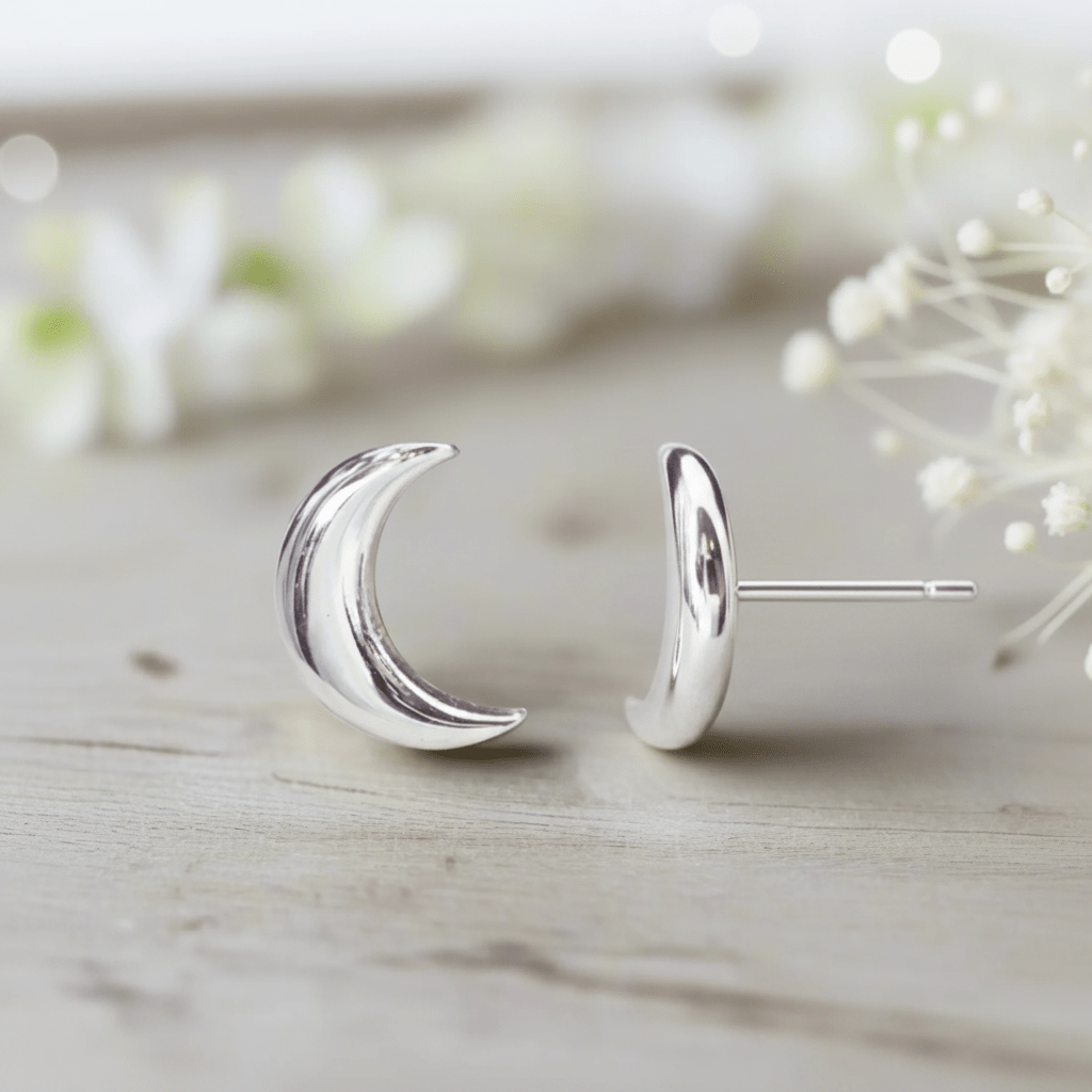 Silver crescent moon stud earrings on a light wooden surface with blurred flowers in the background
