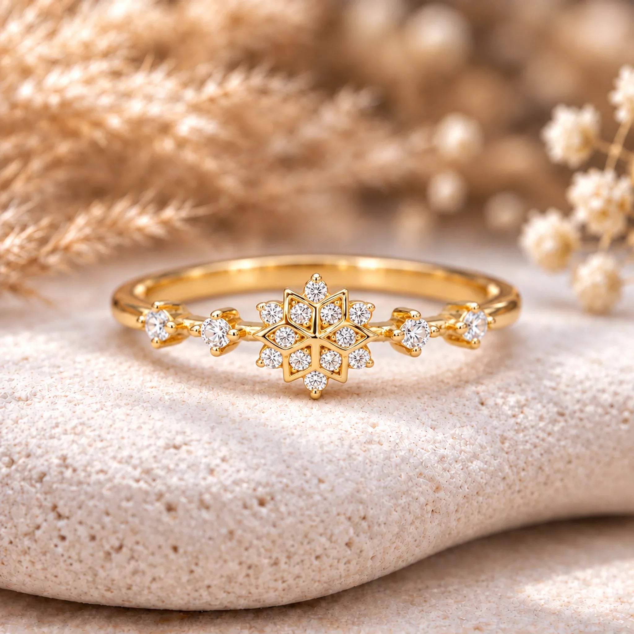 Dainty gold snowflake ring, front view on a pale stone block with softly blurred dried grasses and white flowers
