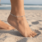 Foot wearing a minimalist sterling silver anklet on a sandy beach with ocean in the background