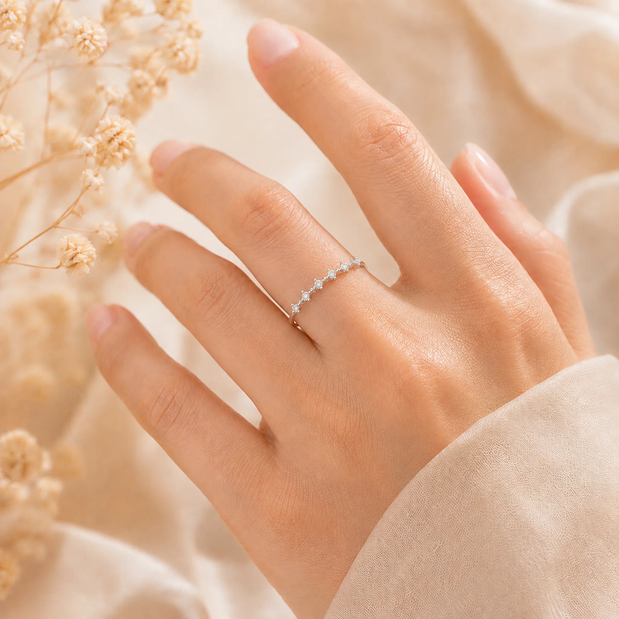 A delicate silver ring with a row of small clear stones worn on a person's ring finger, shown against a soft beige background with dried flowers and a light-colored sleeve.
