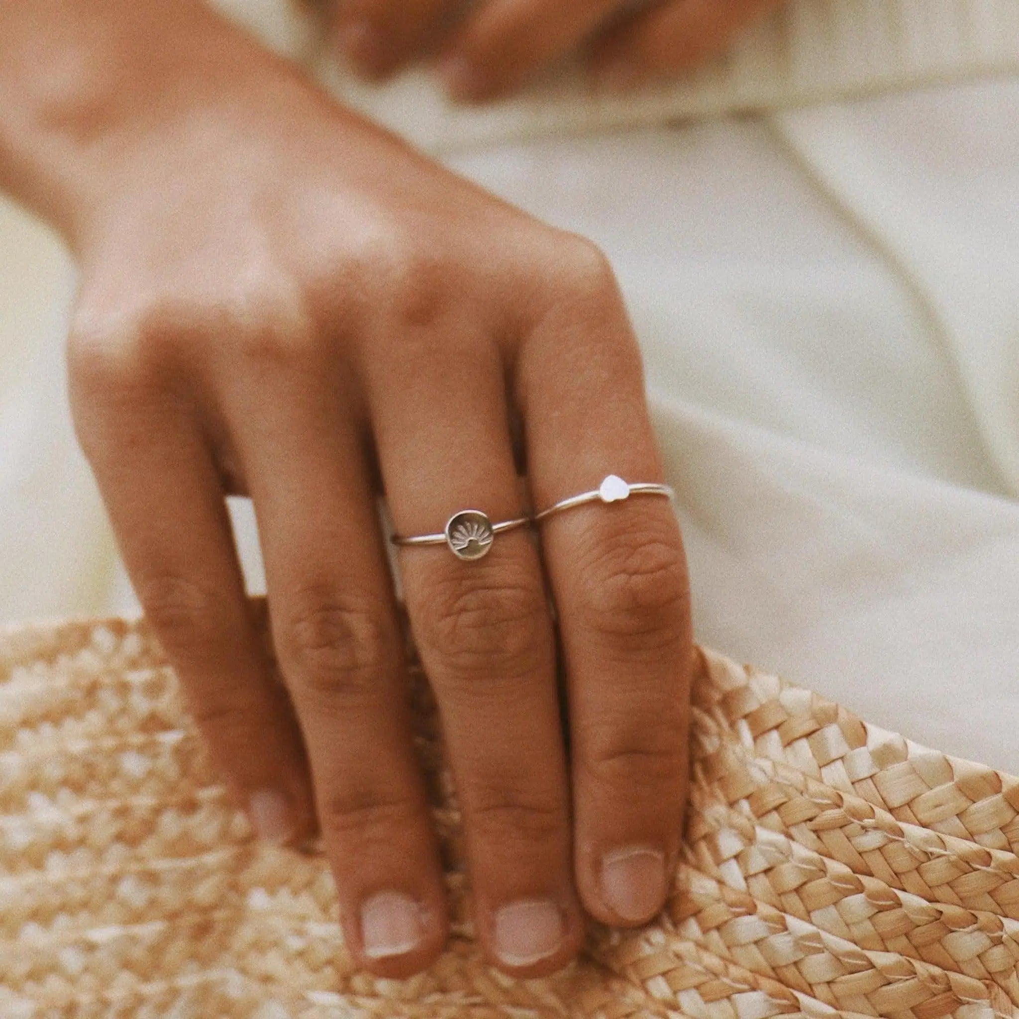A close-up of a hand wearing two delicate silver rings, resting on a woven straw hat with a soft cream-colored garment in the background.