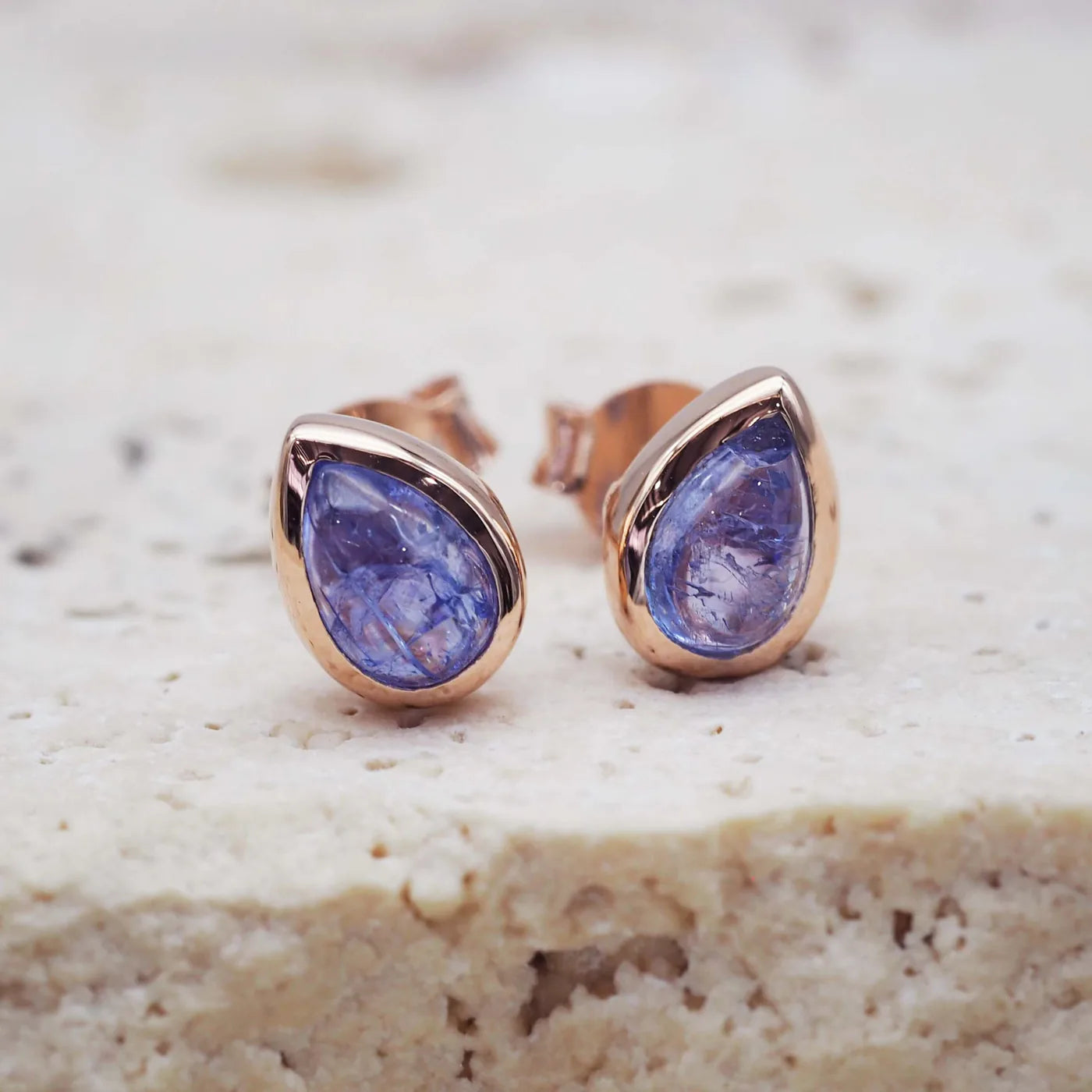 Teardrop-shaped blue gemstone earrings with rose gold settings on a textured beige surface.