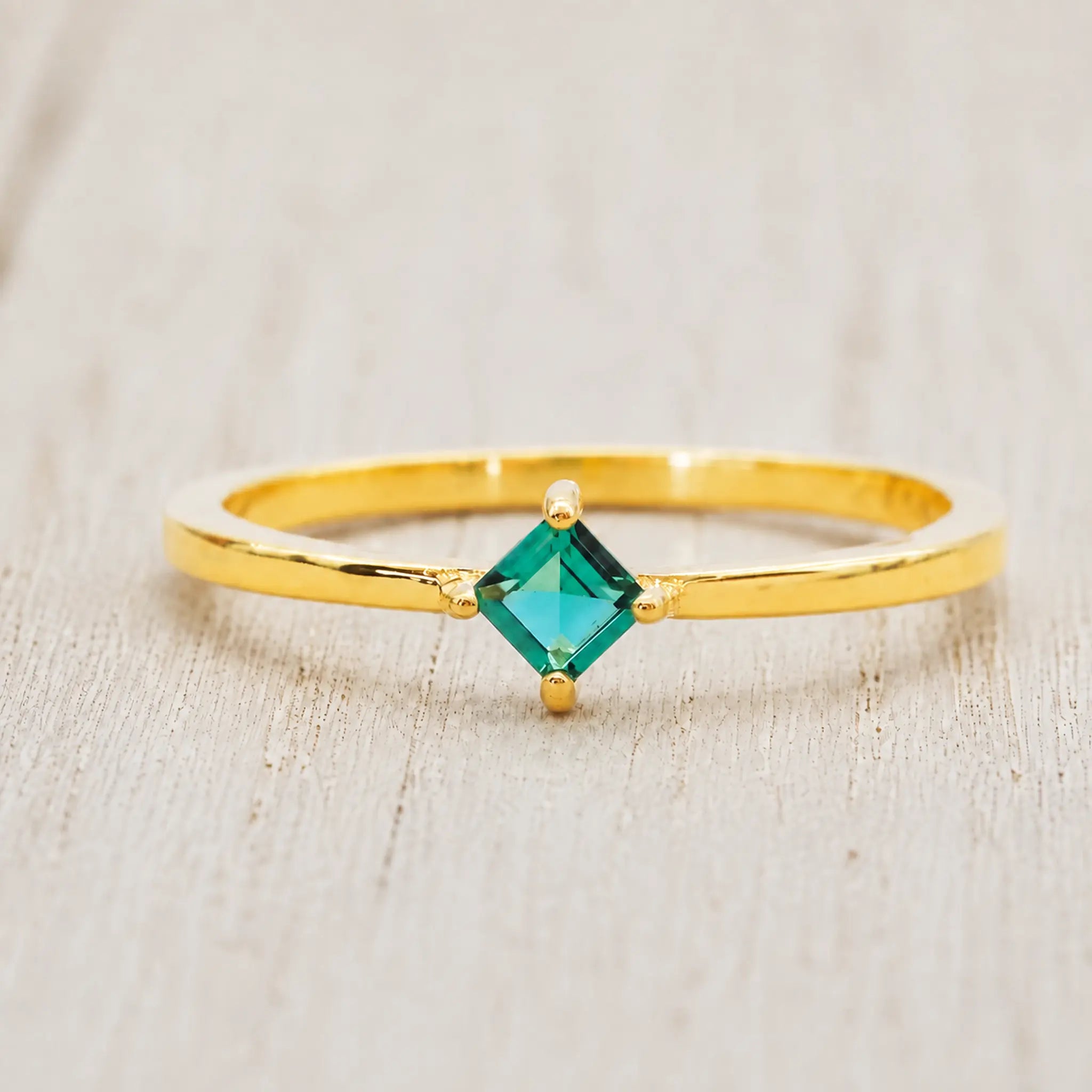 Front view of a gold-plated ring with a princess-cut emerald-green cubic zirconia on pale rustic wood

