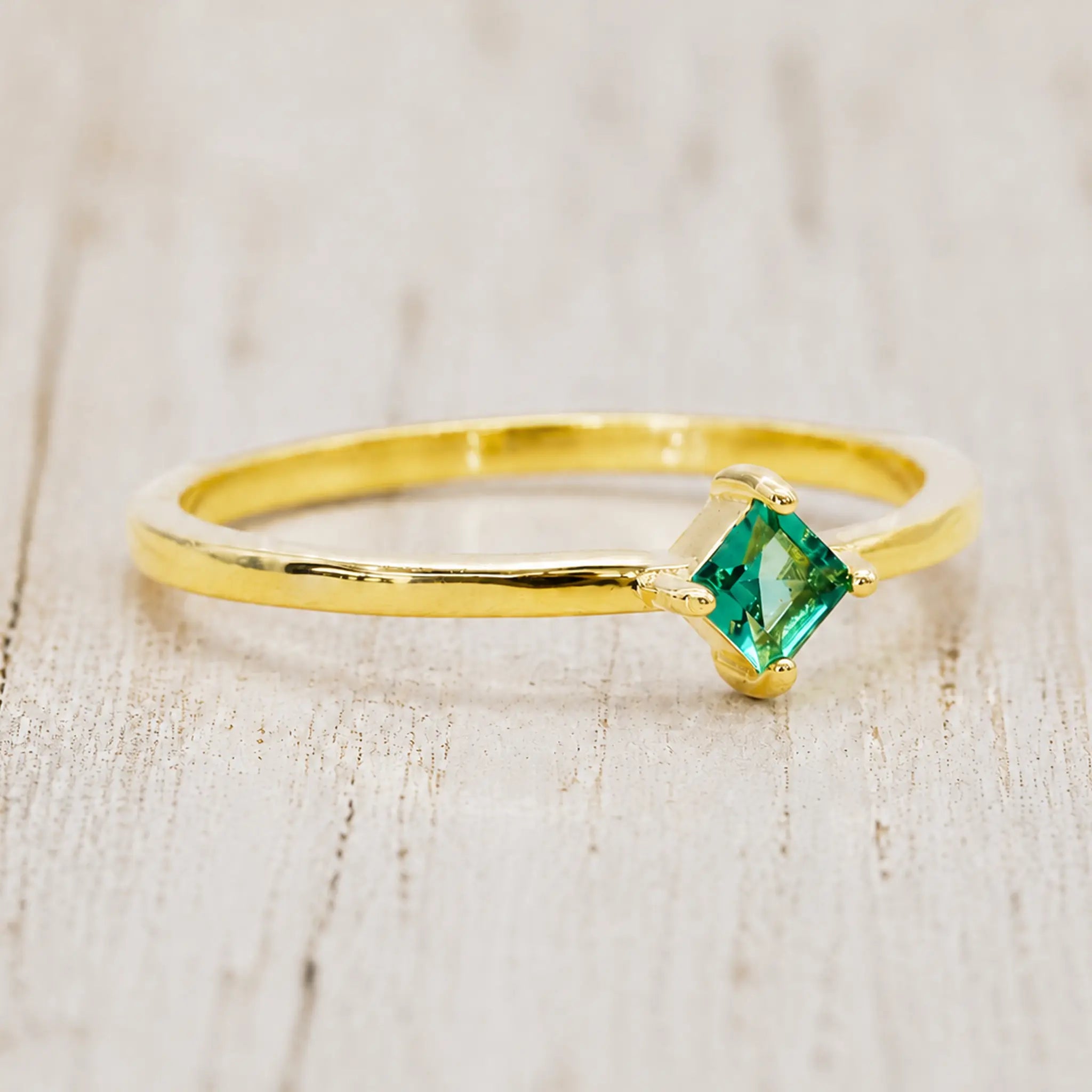 Gold-plated princess-cut emerald-green cubic zirconia ring on a pale rustic wood background
