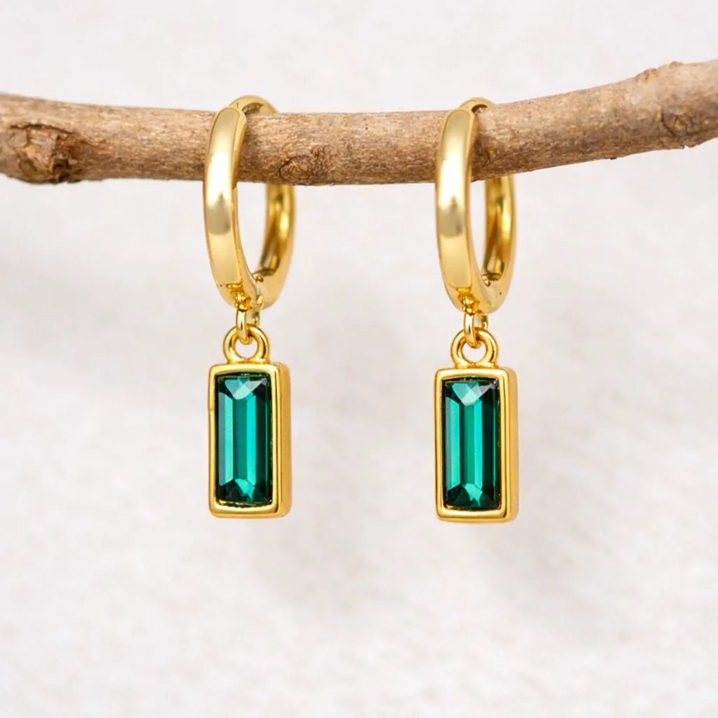 Gold hoop earrings with green gemstones hanging on a branch.