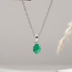 A silver pendant necklace featuring an oval-cut green gemstone, suspended from a delicate silver chain against a soft, light gray background with a subtle dried plant stem on the right.