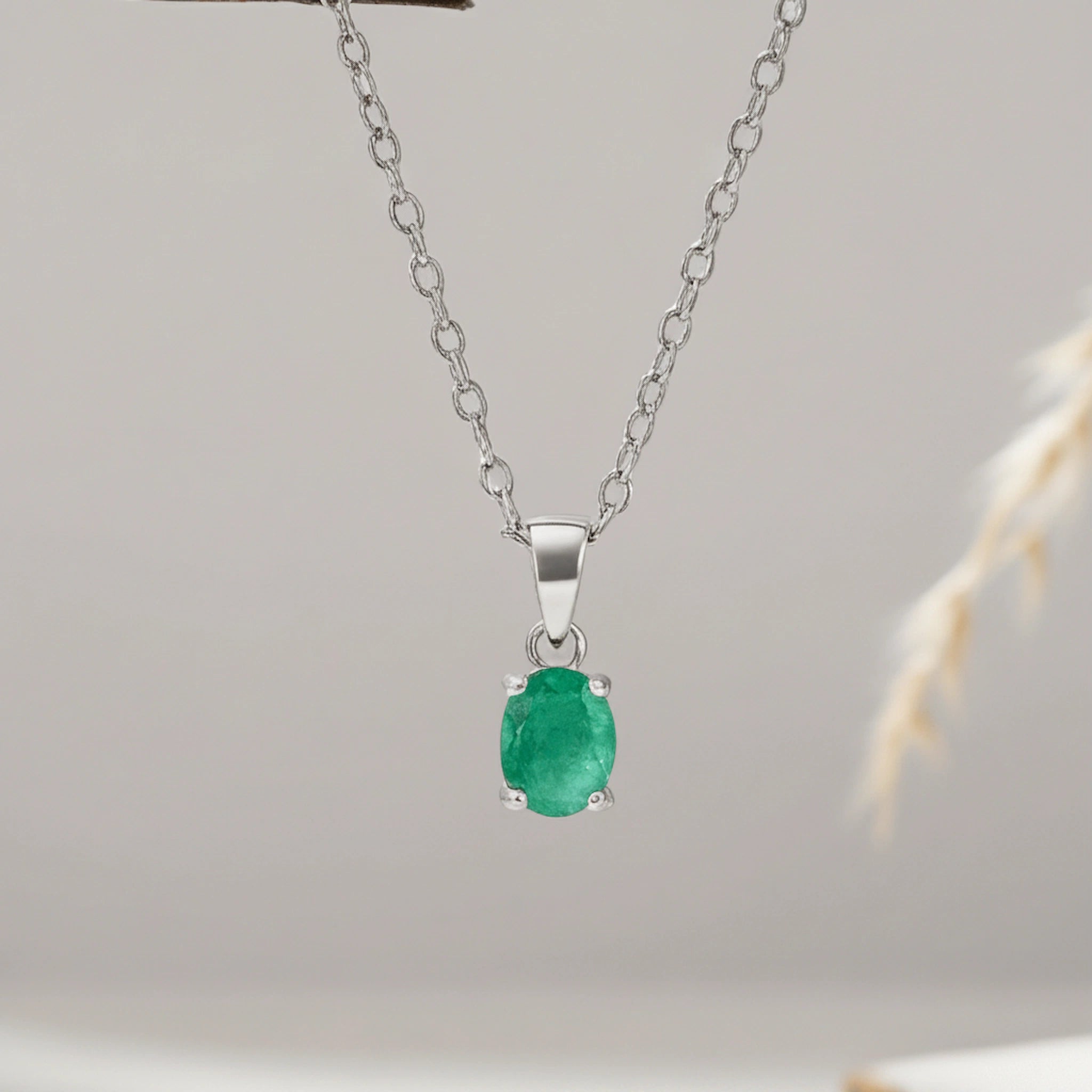 A silver pendant necklace featuring an oval-cut green gemstone, suspended from a delicate silver chain against a soft, light gray background with a subtle dried plant stem on the right.