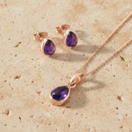 Rose gold jewelry set with purple gemstones on a beige stone surface