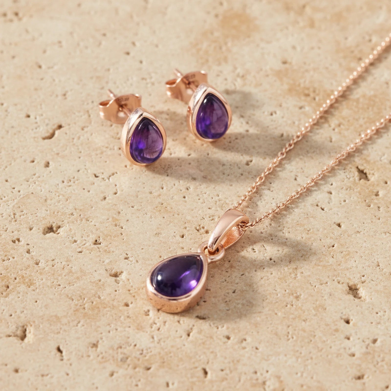 Rose gold jewelry set with purple gemstones on a beige stone surface