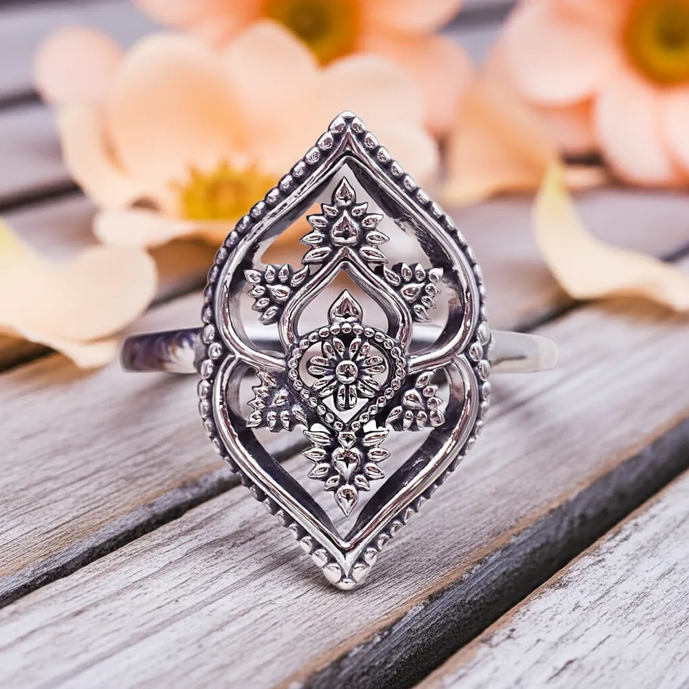 Silver ring with intricate design on a wooden surface with blurred flowers in the background