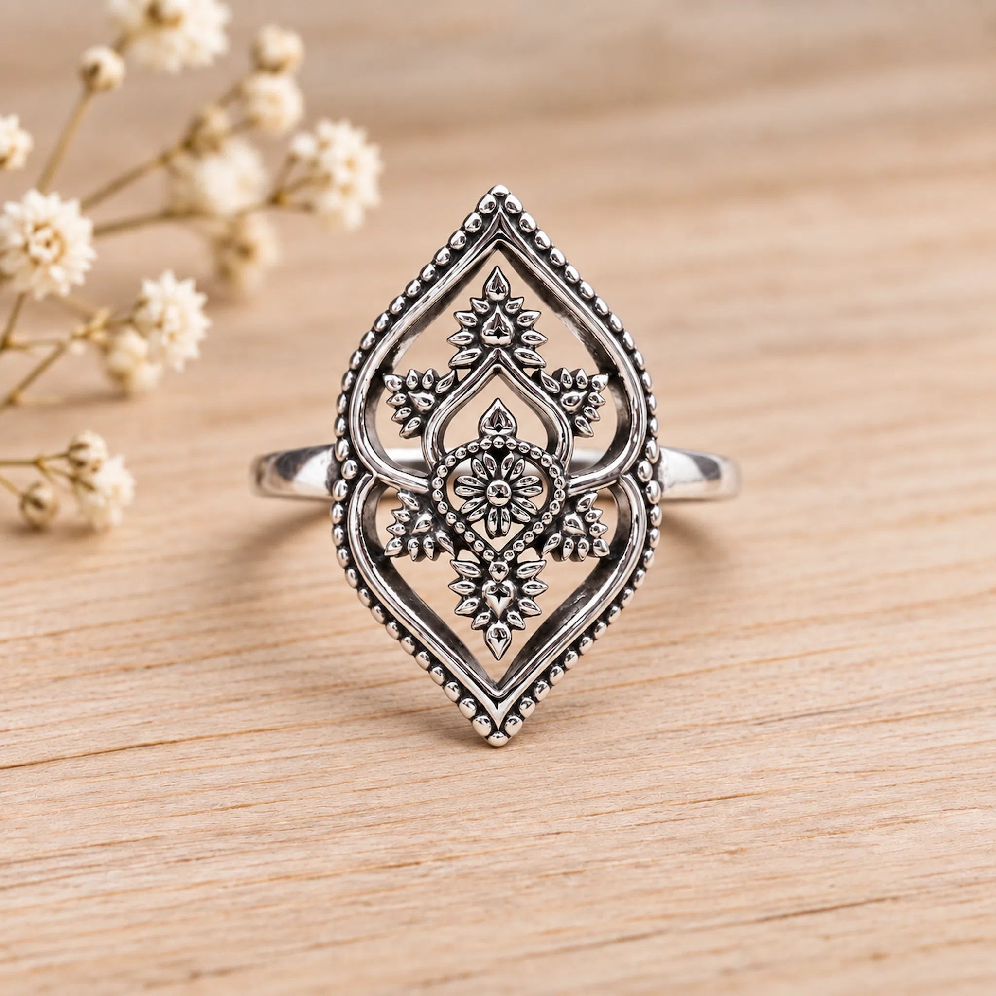 Silver ring with intricate design on a wooden surface with blurred flowers in the background