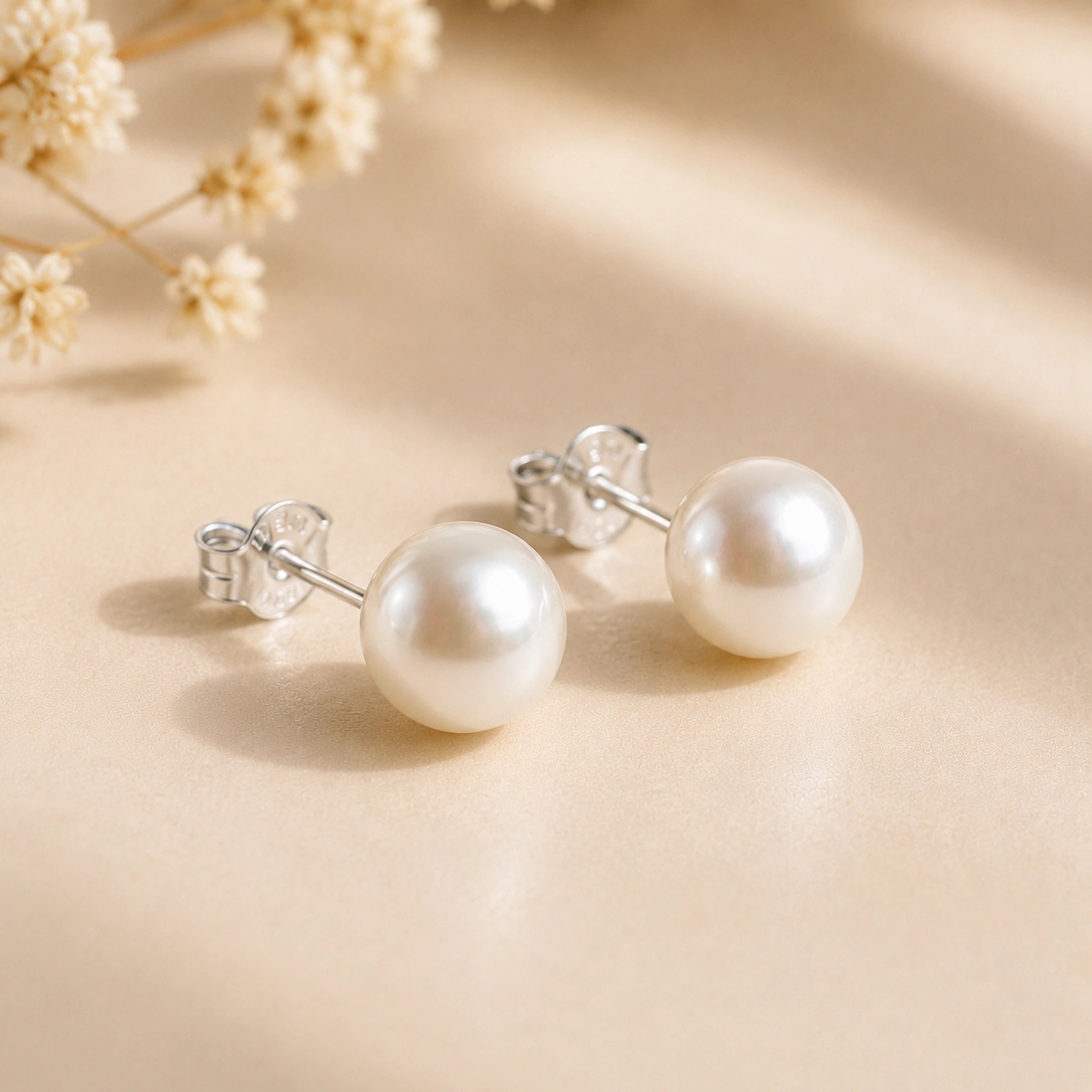 A pair of round white freshwater pearl earrings with silver posts and silver butterfly backings, displayed on a soft beige surface with dried flowers in the background.