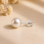 A single freshwater pearl stud earring with a silver post and butterfly back, featuring a sterling silver 925 stamp on the backing. The earring is displayed on a warm beige textured surface with soft shadows and blurred dried flowers in the background.