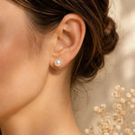 A close-up of a woman's ear wearing a single round white pearl stud earring, with her hair in a low bun and small dried flowers in the softly lit background.