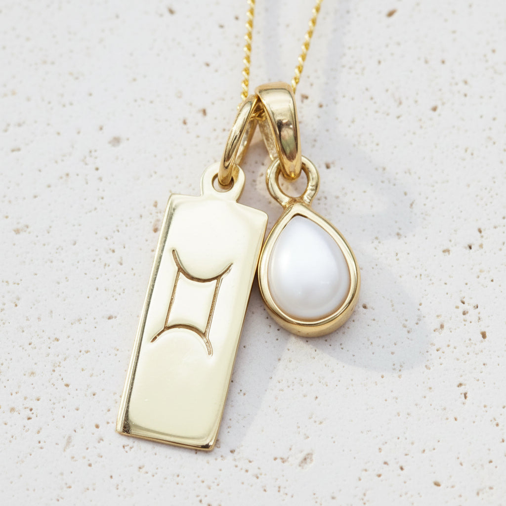 Gold necklace with gemini zodiac pendant and tear drop pearl pendant on a textured background