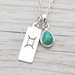Silver necklace with a green teardrop pendant and a rectangular tag with a symbol on a wooden surface.
