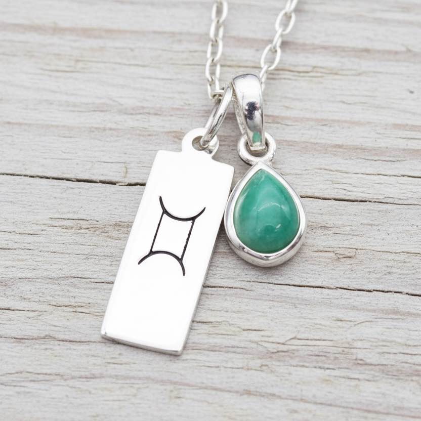 Silver necklace with a green teardrop pendant and a rectangular tag with a symbol on a wooden surface.