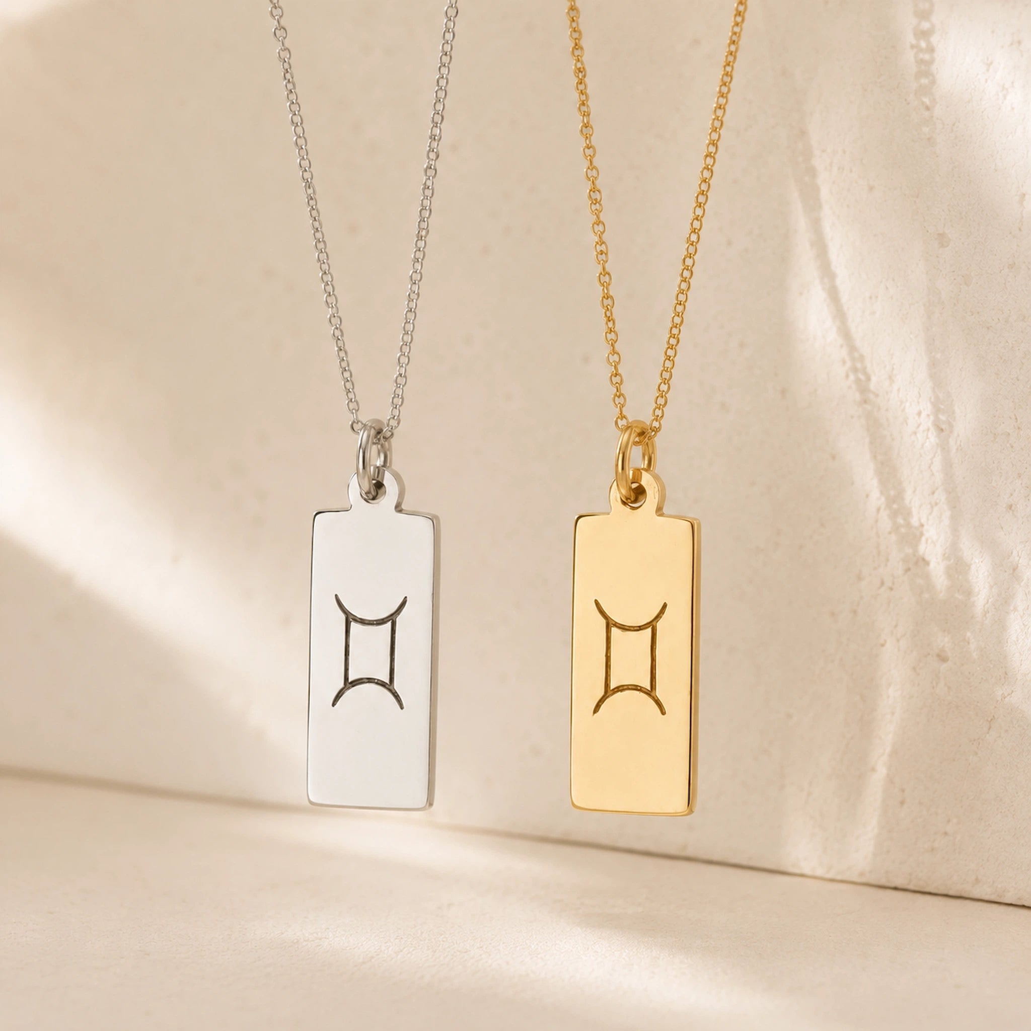gold and sterling silver fine chain necklaces with rectangle pendants engraved with the gemini zodiac line glyph symbol hanging.