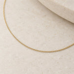 A delicate gold chain necklace draped in a gentle curve across a light beige textured stone surface.