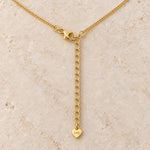 Gold chain necklace with a lobster clasp, an adjustable extension chain, and a small heart-shaped pendant engraved with initials, displayed on a light beige stone surface.