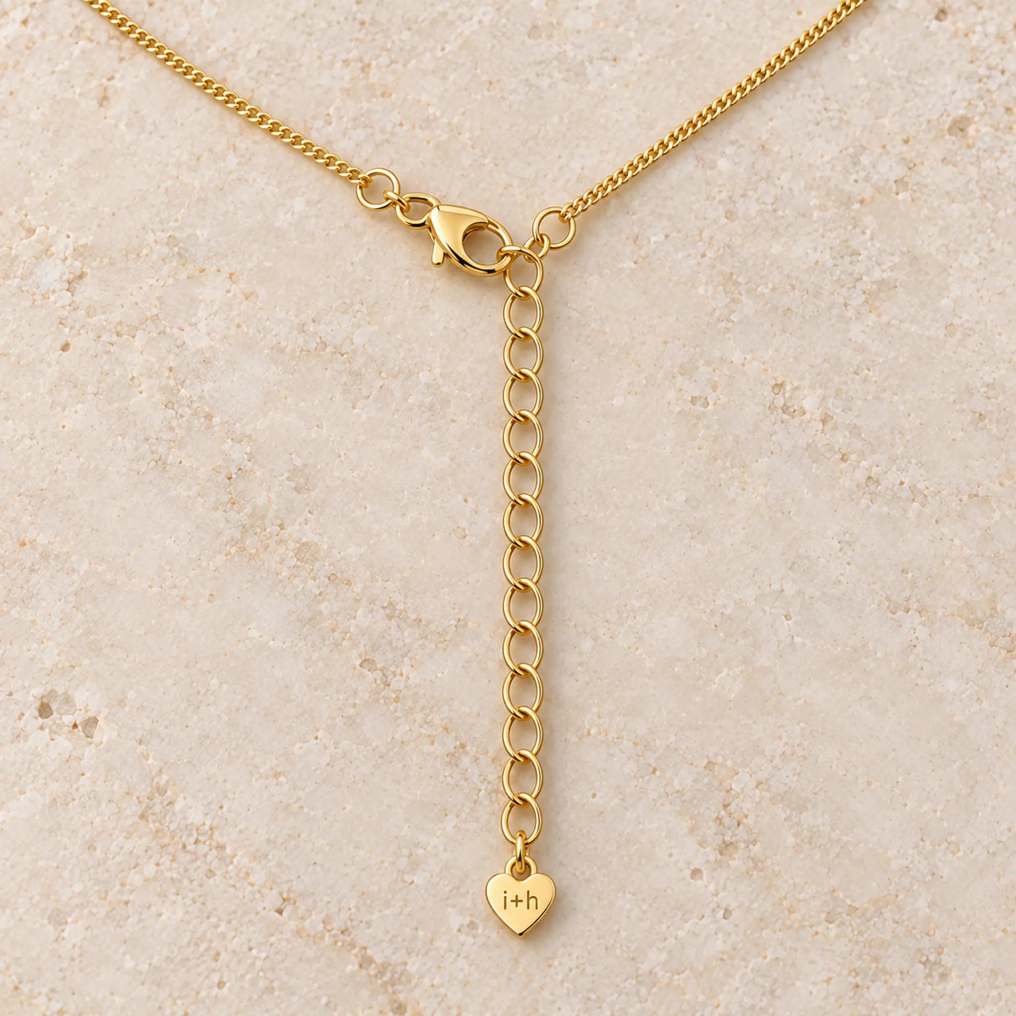 Gold chain necklace with a lobster clasp, an adjustable extension chain, and a small heart-shaped pendant engraved with initials, displayed on a light beige stone surface.