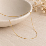A delicate fine 9k gold chain necklace draped over a light beige ceramic bowl on a textured beige surface with dried flowers in the background.