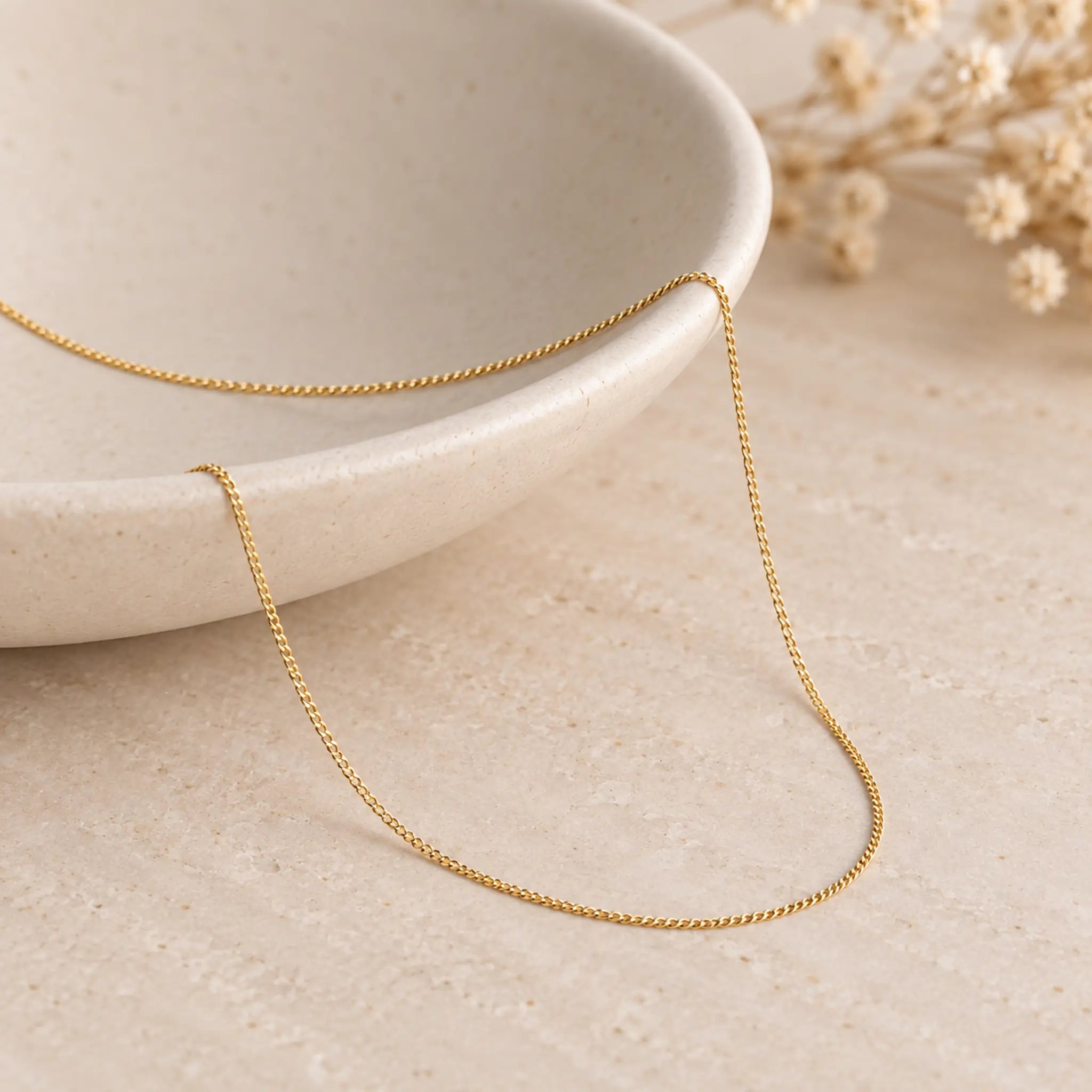A delicate fine 9k gold chain necklace draped over a light beige ceramic bowl on a textured beige surface with dried flowers in the background.