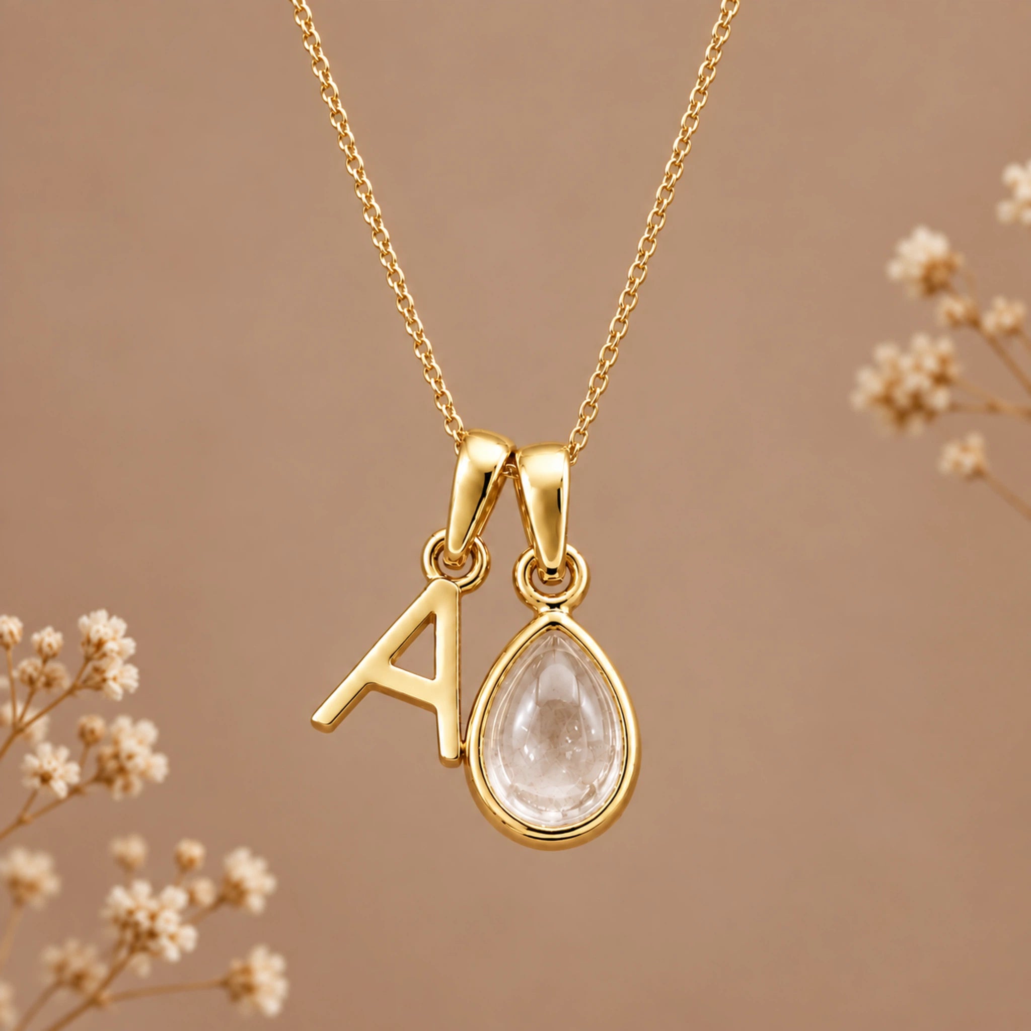 Gold initial necklace with a letter 'A' pendant and a teardrop-shaped clear gemstone pendant, displayed on a warm brown background with dried flowers.