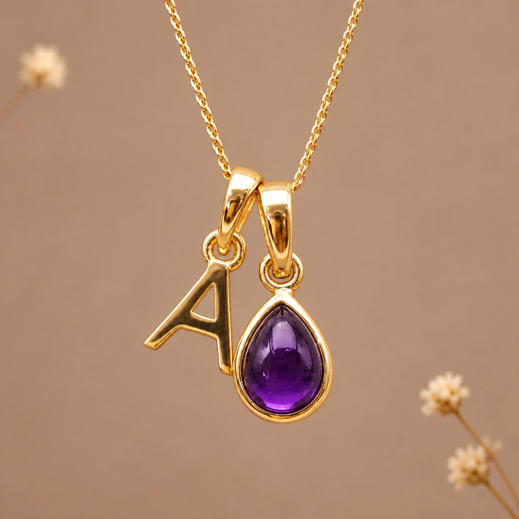 Gold initial pendant necklace featuring the letter 'A' and a february birthstone teardrop-shaped natural amethyst gemstone charm, displayed against a warm beige background with subtle dried flowers.