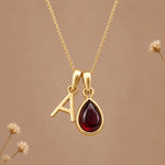 Gold initial pendant necklace featuring the letter 'A' and a teardrop-shaped dark red january birthstone natural garnet gemstone charm, displayed against a warm beige background with subtle dried flowers.