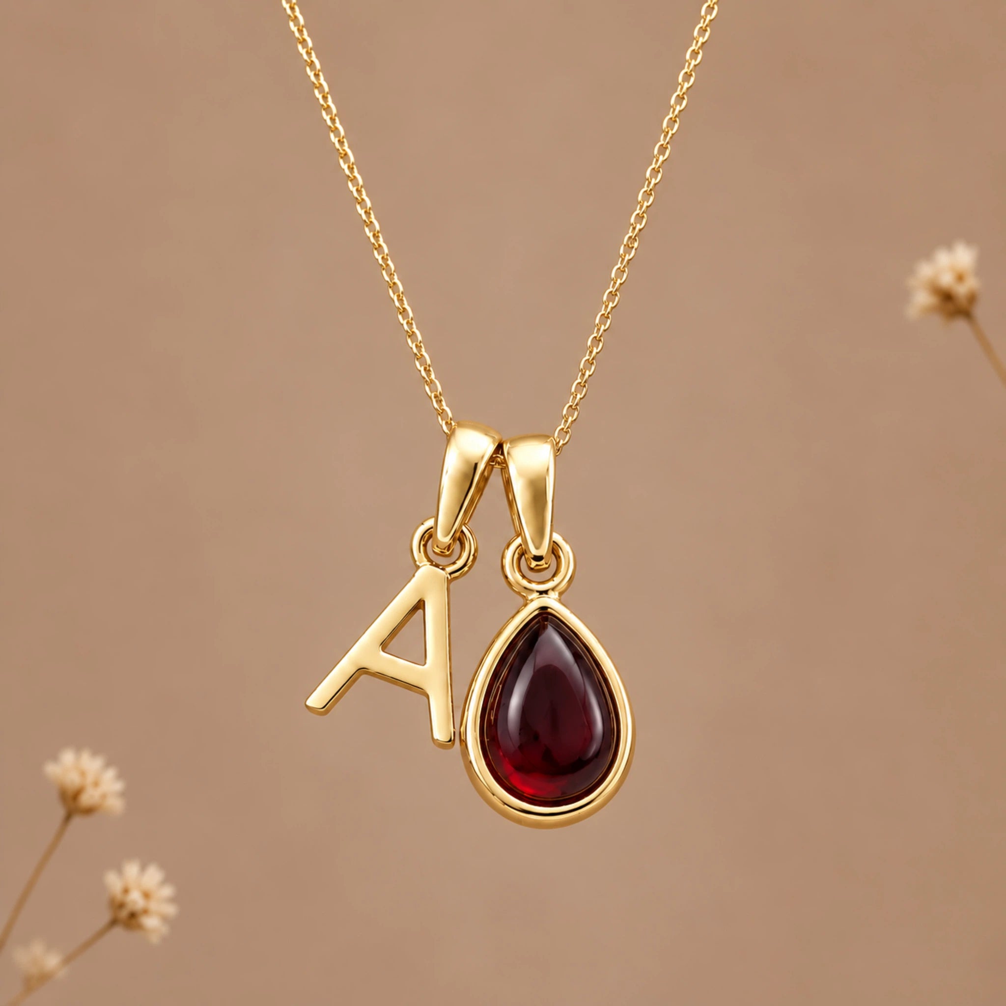 Gold initial pendant necklace featuring the letter 'A' and a teardrop-shaped dark red january birthstone natural garnet gemstone charm, displayed against a warm beige background with subtle dried flowers.