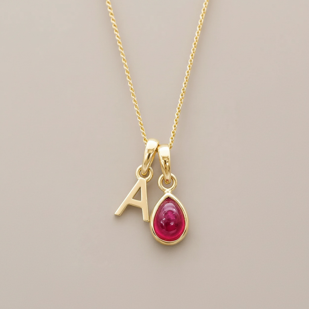 A gold initial necklace featuring a letter 'A' charm and a teardrop-shaped red natural ruby gemstone pendant, displayed on a neutral beige background.