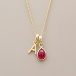 A gold initial necklace featuring a letter 'A' charm and a teardrop-shaped red natural ruby gemstone pendant, displayed on a neutral beige background.