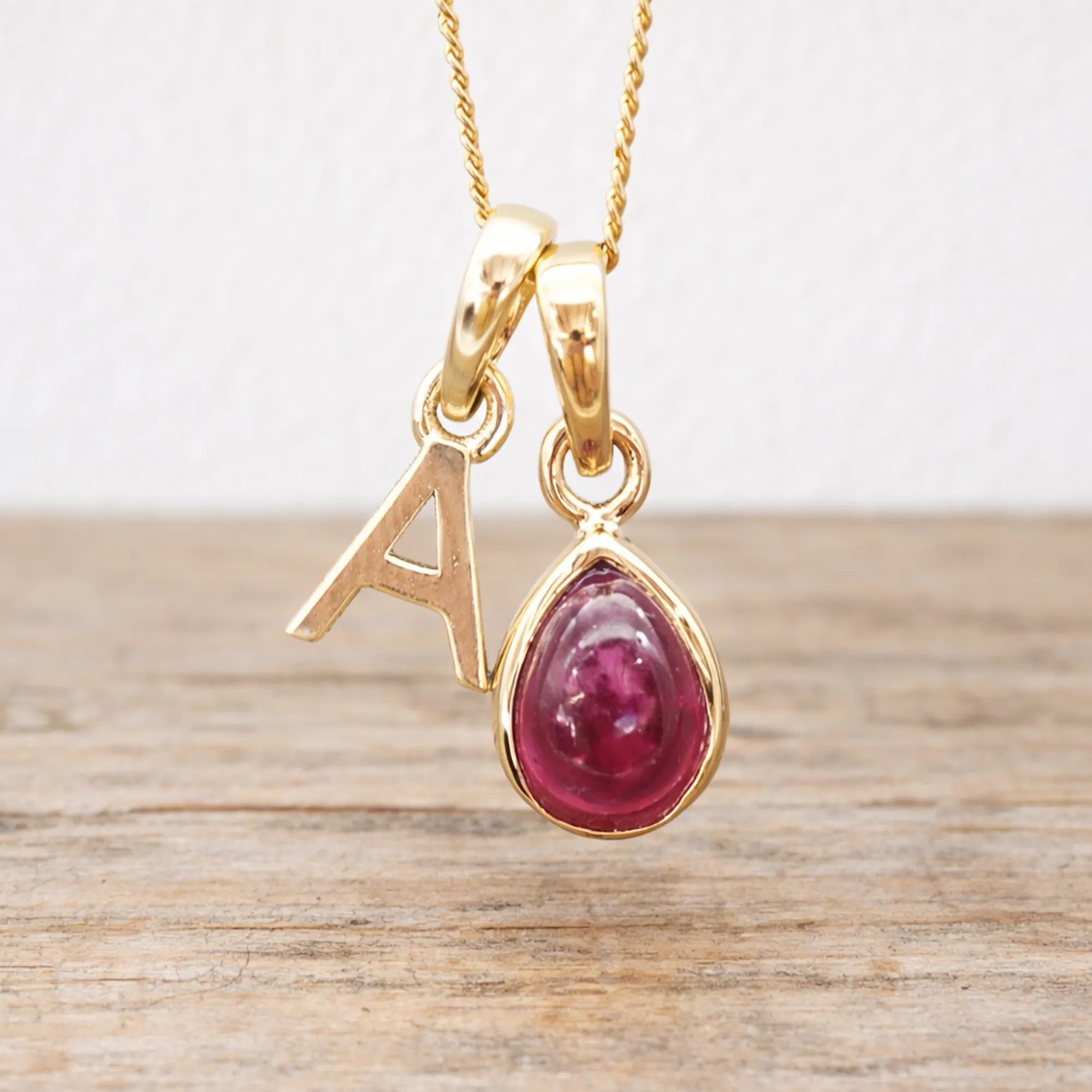 Gold necklace with a teardrop-shaped ruby pendant and letter 'A' charm on a wooden surface.